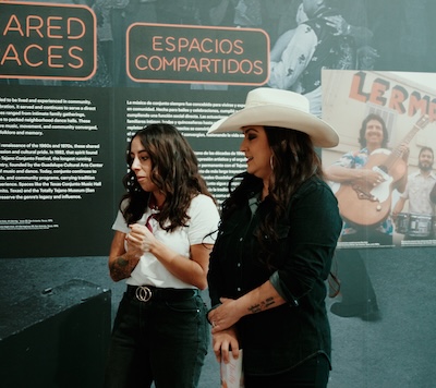 ITC Head Curator Bianca Alvarez leads Shelly Lares on a tour of “Puro Conjunto: Border(less) Sound.” Alvarez explains a display, off-camera, of German musical instruments.